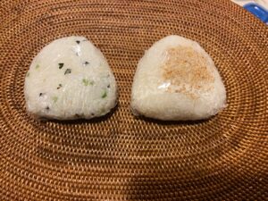 Two triangular rice balls wrapped in plastic, one topped with sesame seeds, and the other wrapped in seaweed.