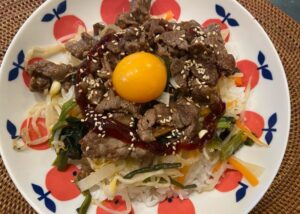 A colorful bibimbap bowl with vegetables, grilled beef, and a raw egg yolk on a white plate with apple patterns.