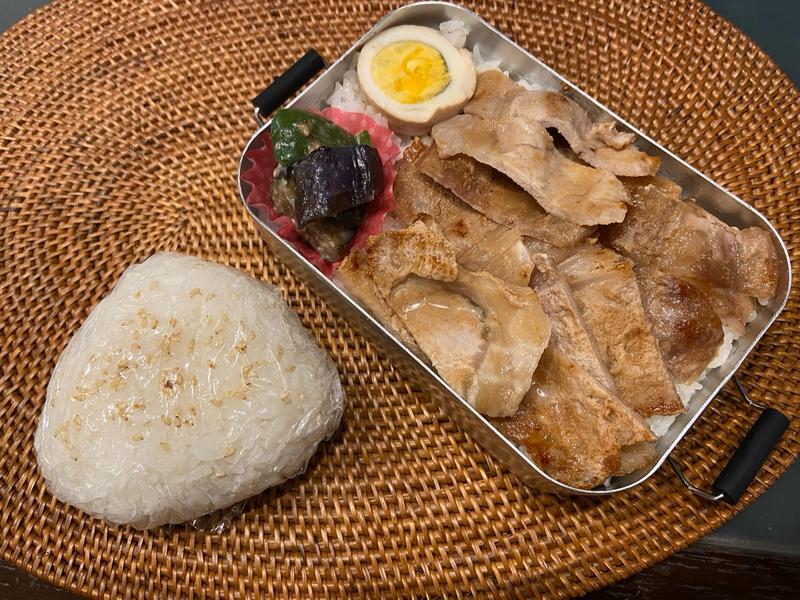 Grilled beef bento box with boiled egg, vegetables, and a sesame rice ball on a woven placemat.