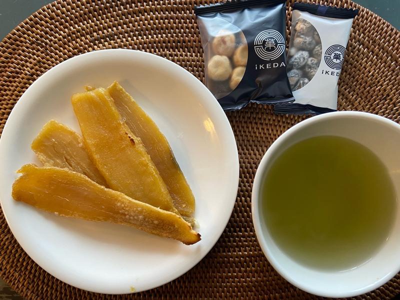 Dried sweet potato slices on a plate, green tea in a cup, and Ikeda bean snacks on a woven placemat.