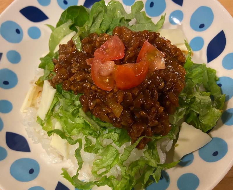 A close-up of homemade taco rice with seasoned minced meat, fresh lettuce, cherry tomatoes, and cheese on a blue patterned plate.