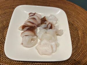 Octopus sashimi slices on a white square plate with a woven placemat underneath.