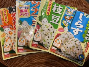 Four packages of Japanese onigiri mix in salmon, tuna mayo, edamame, and shirasu flavors, placed on a woven placemat.