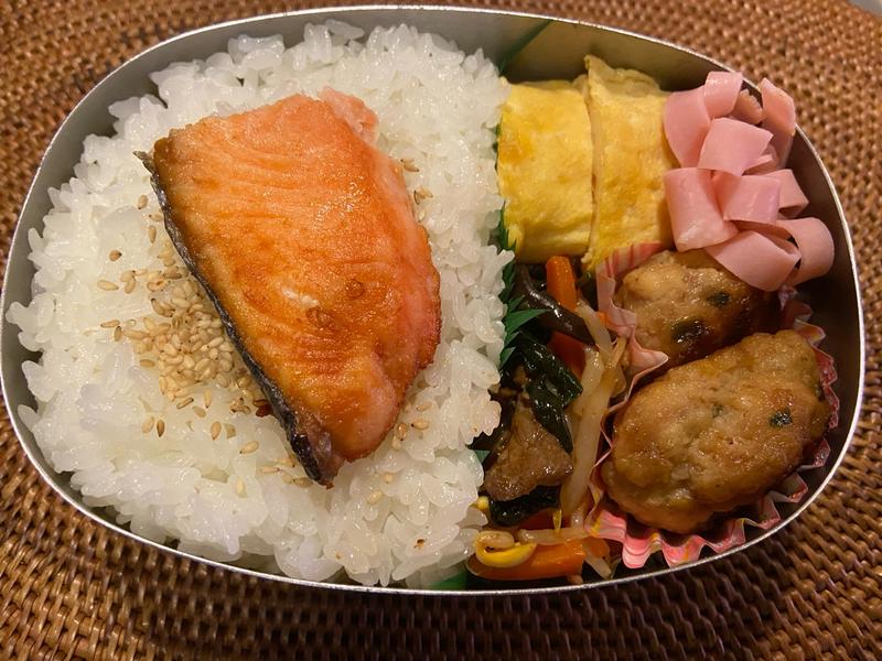 apanese bento with grilled salmon, tamagoyaki, chicken meatballs, and vegetables served in a metal box on a woven placemat