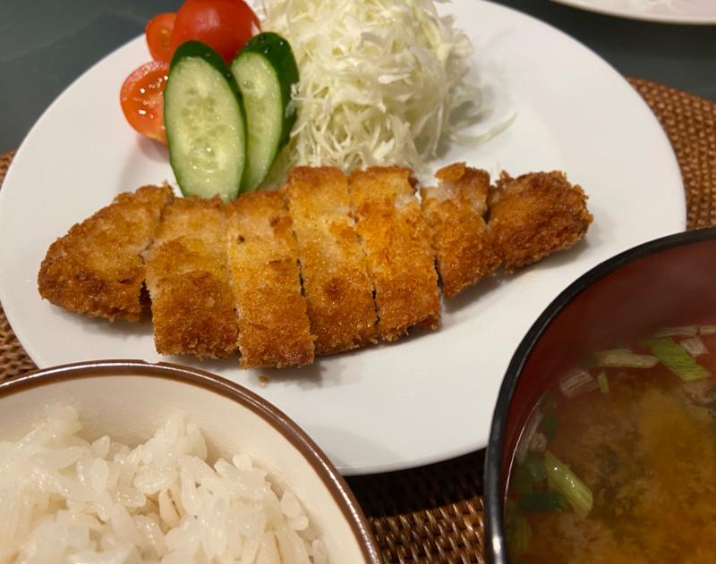 A plate of golden-brown tonkatsu with shredded cabbage, sliced cucumbers, cherry tomatoes, steamed rice, and miso soup.